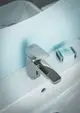 Chromarmatur mit Sensorsteuerung, montiert über einem Waschbecken. Ideal Standard Marke. Im Hintergrund sind Pflegeprodukte auf einem weißen Ablageflächen sichtbar.